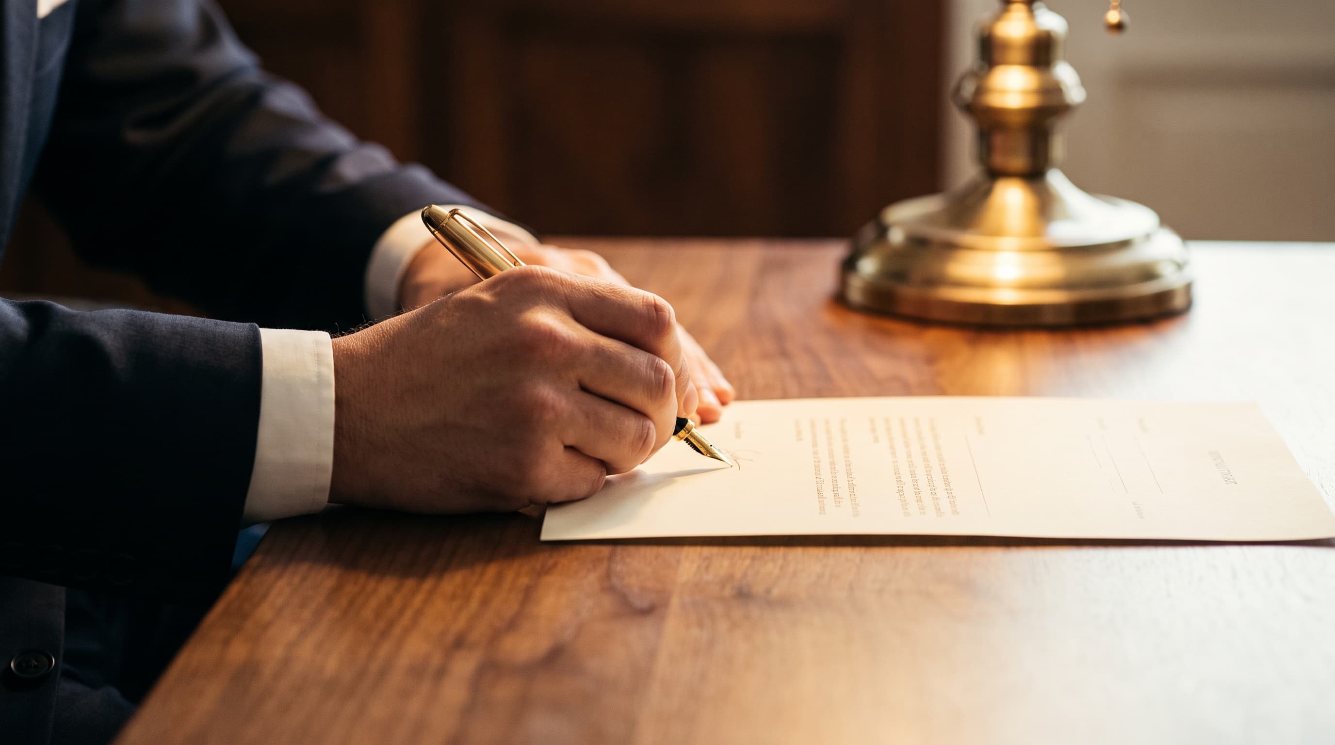 Mano firmando un documento con pluma sobre un escritorio de nogal